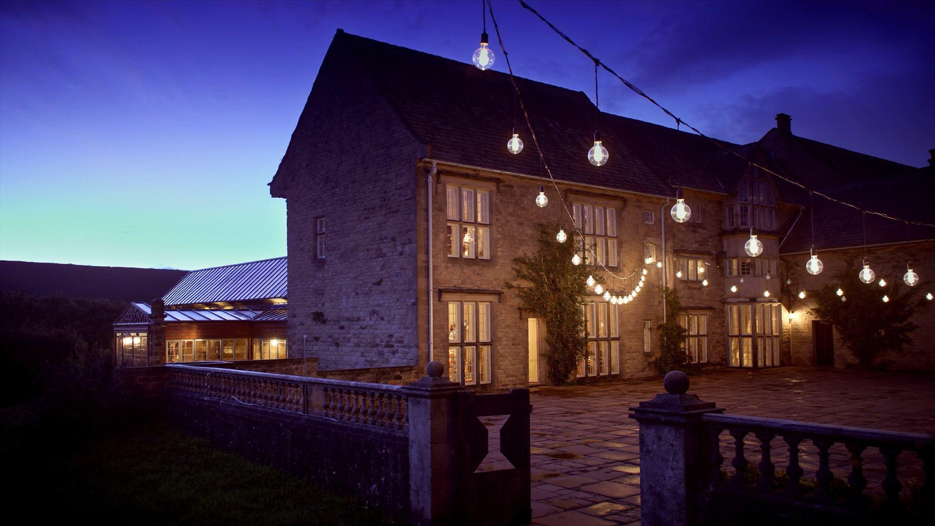The hall at dusk across the courtyard, windows glowing warm, festoon bulbs strung overhead against a deep blue sky