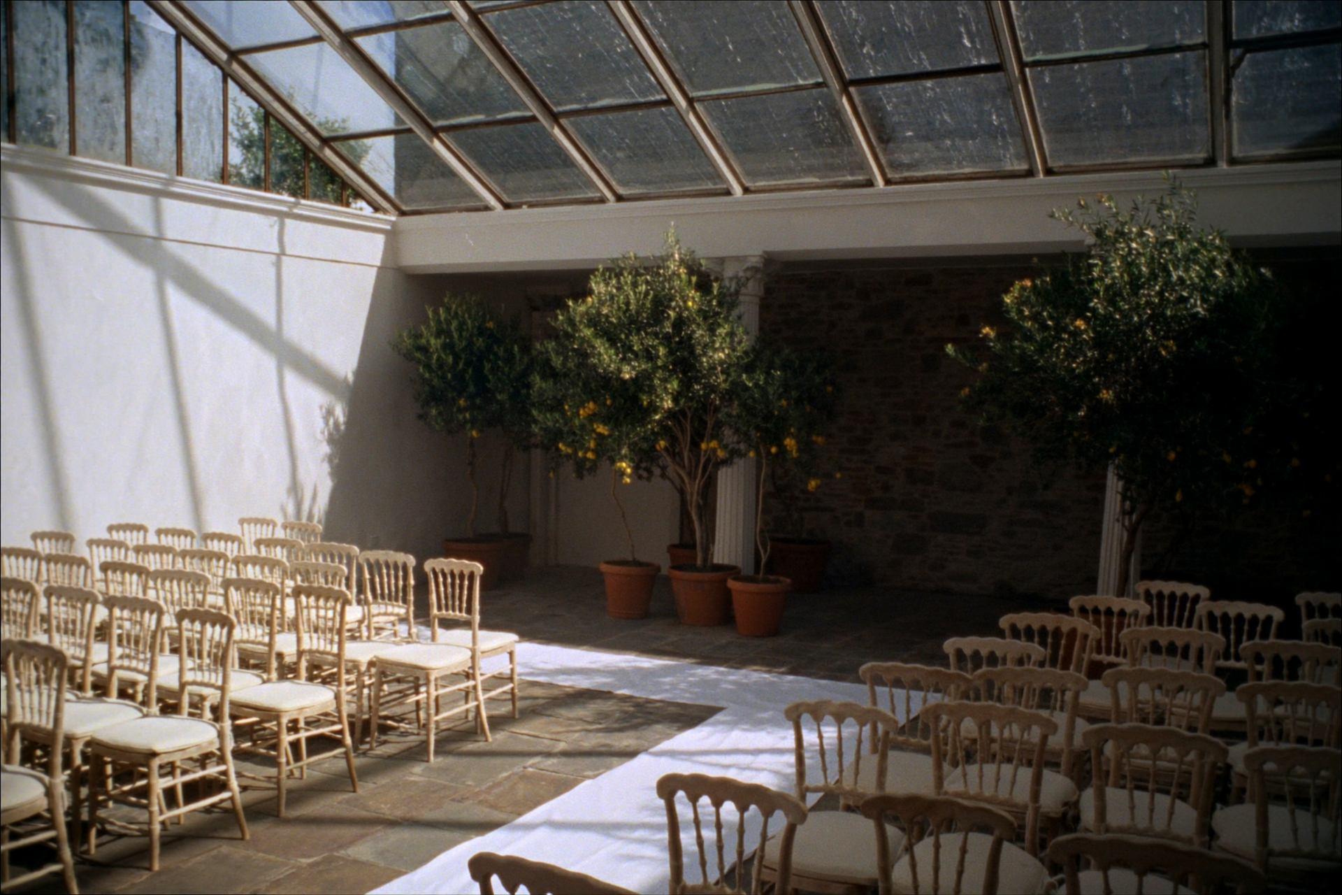 The glass orangery set with pale chairs either side of a stone aisle, morning sun falling through the glazed roof